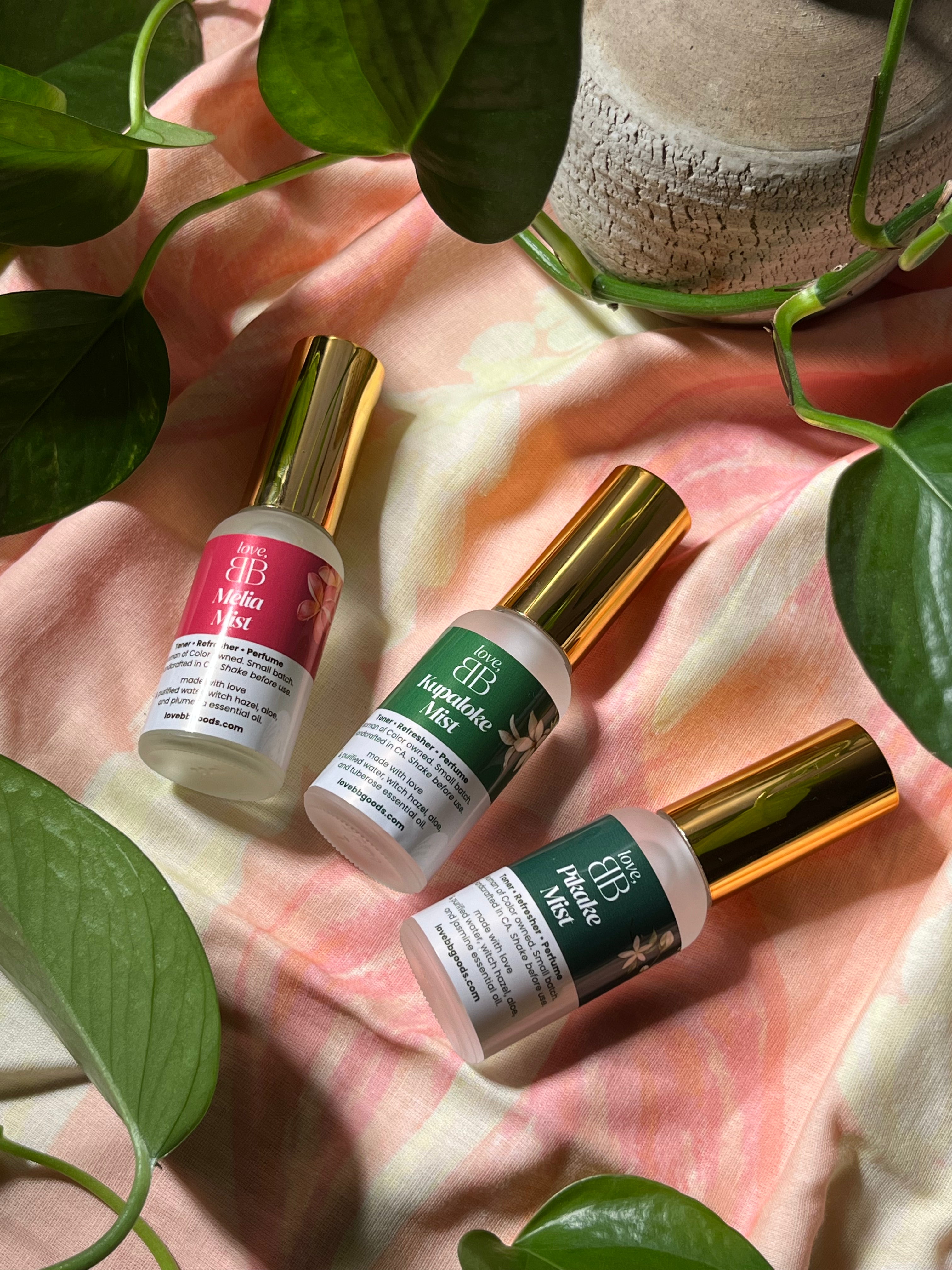 Three small bottles with gold caps on a pink fabric background with green leaves.